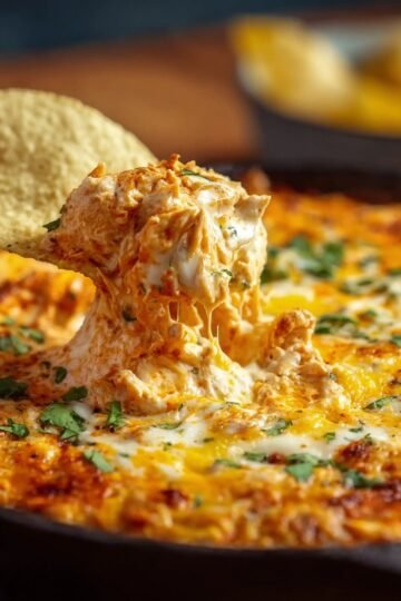 buffalo chicken dip Tortilla chip being dipped into creamy buffalo chicken dip with melted cheese and herbs in a skillet.