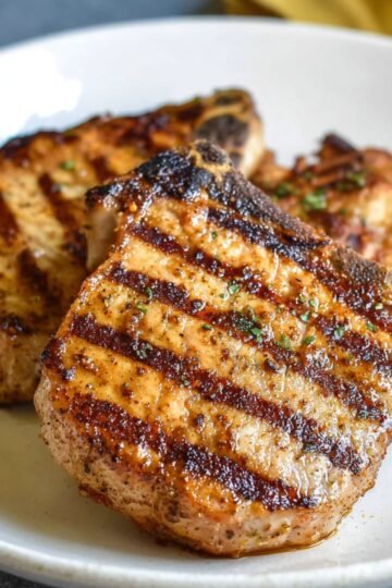 Pork Chop Marinade A plate with two golden, grilled pork chops that have perfect grill marks. The pork is tender and juicy, topped with a light seasoning, ready to be served with a side dish.
