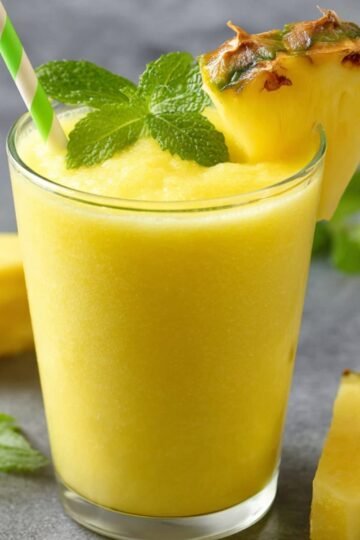 pineapple smoothie A glass of pineapple smoothie with a green striped straw, garnished with a slice of pineapple and a mint sprig, placed on a gray surfac