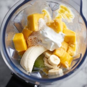 A close-up view of a blender with frozen mango, banana, yogurt, and other ingredients blending into a smooth mixture.