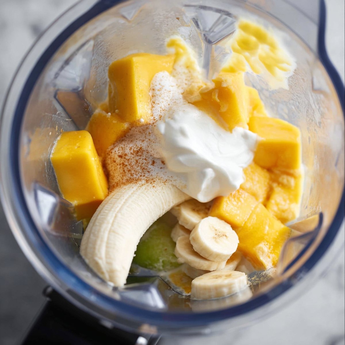 A close-up view of a blender with frozen mango, banana, yogurt, and other ingredients blending into a smooth mixture.