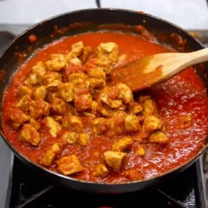 A wooden spoon stirring chicken pieces in a rich, red tomato sauce on a stove. The chicken is cooked and simmering in the sauce.