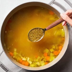 Hand holding a spoon of seasoning over a pot of chicken broth with carrots and celery
