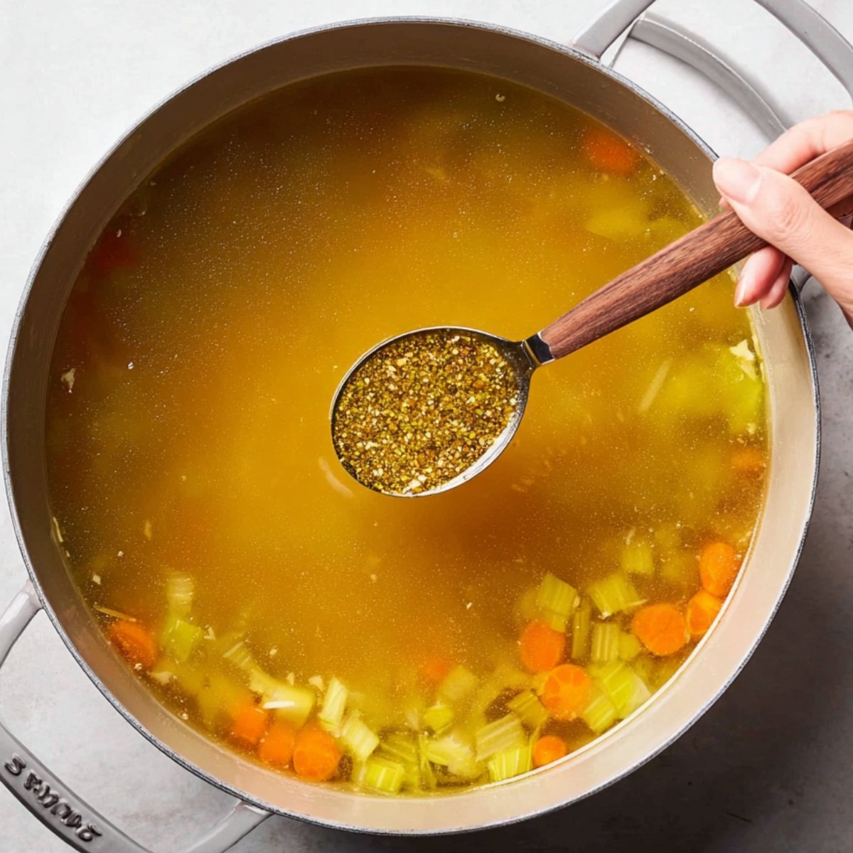 Hand holding a spoon of seasoning over a pot of chicken broth with carrots and celery