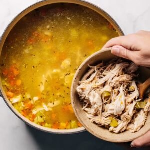 Shredded cooked chicken being added to a pot of chicken soup with vegetables