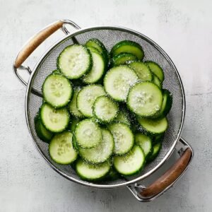 Sliced cucumbers sprinkled with salt in a metal colander for draining excess moisture