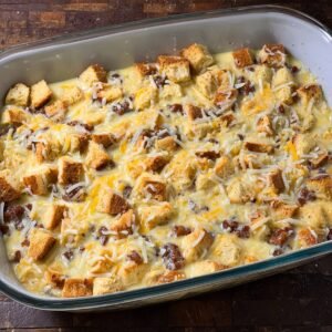 A baking dish filled with a mixture of bread cubes, cooked sausage, and shredded cheese, all soaked in an egg mixture, ready to be baked into a casserole.