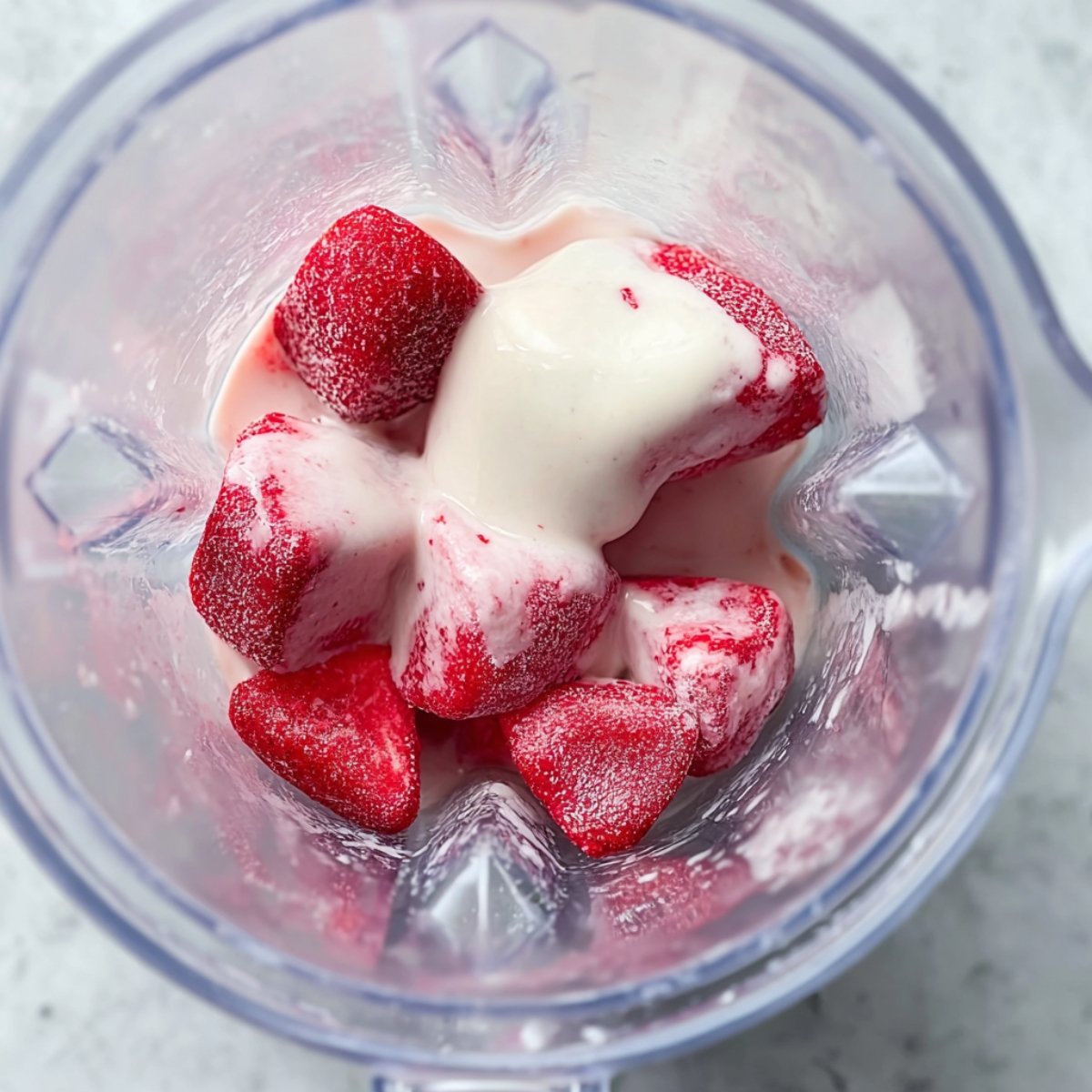Close-up of frozen strawberries and yogurt in a blender, with a creamy texture starting to blend together for a strawberry smoothie.