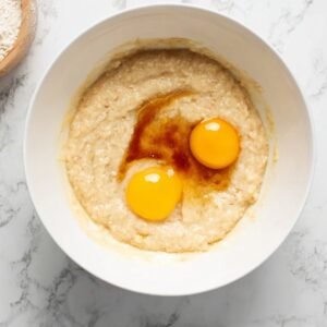 Mashed bananas in a white bowl with two raw eggs and vanilla extract added.