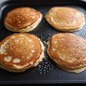 Golden-brown pancakes cooking on a griddle. The pancakes have a slightly crispy edge, with small bubbles on the surface, indicating they are nearly ready to be flipped.