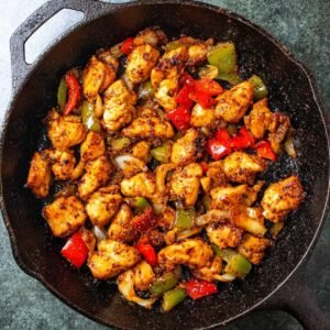 Cooked chicken pieces sautéed with red and green peppers in a cast iron skillet, seasoned and lightly browned.