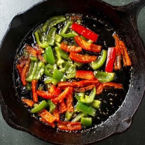 Sliced red and green peppers sautéing in oil in a cast iron skillet, viewed from above.