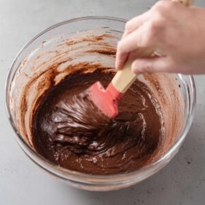 A close-up shot of a spatula stirring thick, glossy brownie batter in a large glass mixing bowl. The rich, chocolate mixture swirls together as it’s being blended by hand, showcasing the texture of the batter."