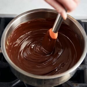A hand gently stirring melted chocolate in a bowl placed over a stove, the chocolate turning silky smooth as it melts. The focus is on the smoothness and consistency of the chocolate, ready to be combined into the brownie mix.