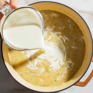 Heavy cream being poured into a pot of potato soup during cooking, creating a rich and creamy texture.