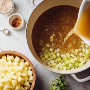 Chicken broth being poured into a pot with sautéed onions and celery, with diced potatoes and seasonings arranged nearby.