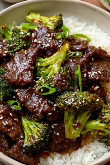 Beef and Broccoli A bowl of beef and broccoli stir-fry served over white rice, with tender beef pieces coated in a glossy sauce and bright green broccoli florets. The dish is garnished with sesame seeds and accompanied by chopsticks, with small bowls of sauce and sliced green onions in the background