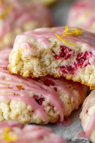 Lemon Raspberry Cookies A close-up of freshly baked lemon raspberry cookies, drizzled with pink glaze and garnished with lemon zest. Some cookies have bites taken out, revealing the soft inside with raspberry chunks