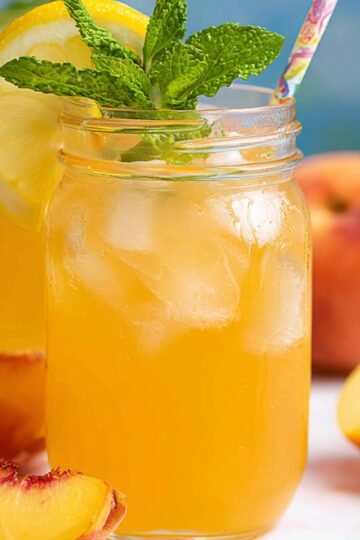 Peach Lemonade A mason jar of peach lemonade garnished with a lemon slice and fresh mint, with a straw, alongside fresh peach slices, creating a summery, refreshing scene.