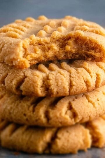 Peanut Butter Cookies A close-up stack of peanut butter cookies, golden brown with fork-pressed ridges; the top cookie is broken in half, showing a soft, crumbly interior against a neutral background.