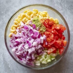 Chopped red onions, tomatoes, and other fresh vegetables are added to a bowl of macaroni, bringing color and texture to the dish.
