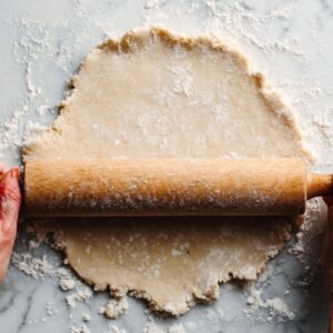 Hands using a rolling pin to flatten pie dough on a marble surface, dusted with flour, preparing the dough for the perfect pie crust.