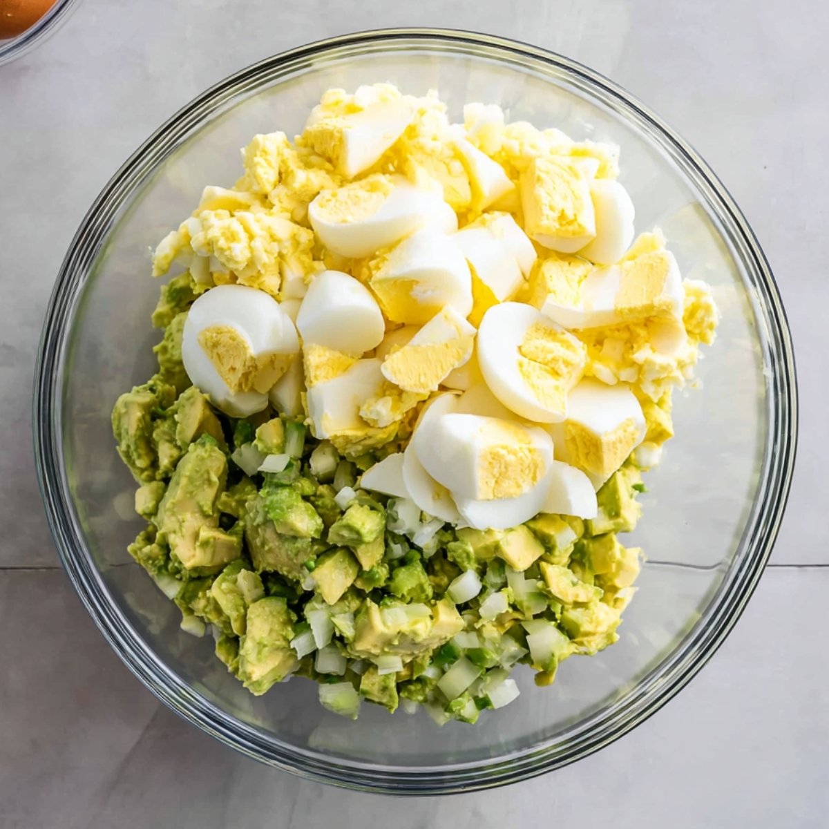 Hard-boiled eggs chopped into small pieces, added to the diced avocado in a mixing bowl, forming the base of a creamy avocado egg salad.
