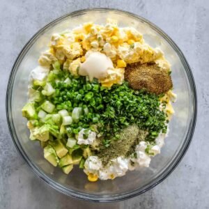 A mixing bowl filled with chopped eggs, diced avocado, green onions, mayonnaise, mustard, fresh herbs, and seasoning, all ready to be mixed into a delicious egg salad.