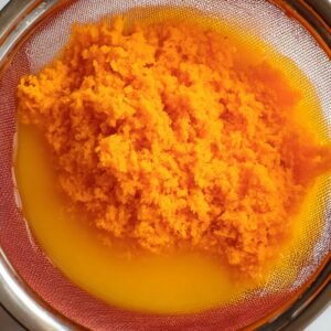 Close-up of freshly strained orange carrot pulp in a sieve, with vibrant orange juice underneath. The texture of the pulp is fluffy and fine, while the juice is a bright, smooth liquid