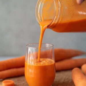 A hand pouring freshly made, vibrant orange Caribbean carrot juice from a jar into a glass. The juice is flowing smoothly with a deep orange color, set against a backdrop of whole carrots.