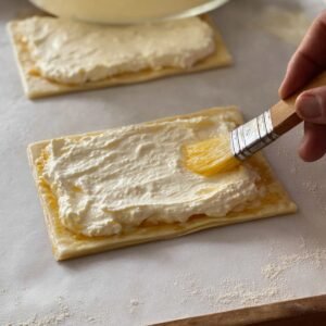 "Spread the creamy cheese filling evenly over each puff pastry square, leaving a small border around the edges. Once the filling is in place, gently brush the edges of the pastry with a beaten egg wash. This will help create a golden, glossy finish once baked. The filling will provide a rich, creamy center, while the egg wash gives the pastry its beautiful color and texture."