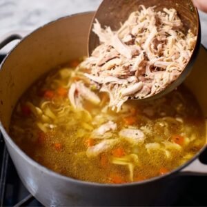 Shredded chicken being added to a pot of simmering soup, making the chicken noodle soup hearty and filling.