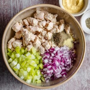 A mixing bowl filled with the ingredients for chicken salad: shredded chicken, diced celery, red onion, mayonnaise, mustard, and seasonings.