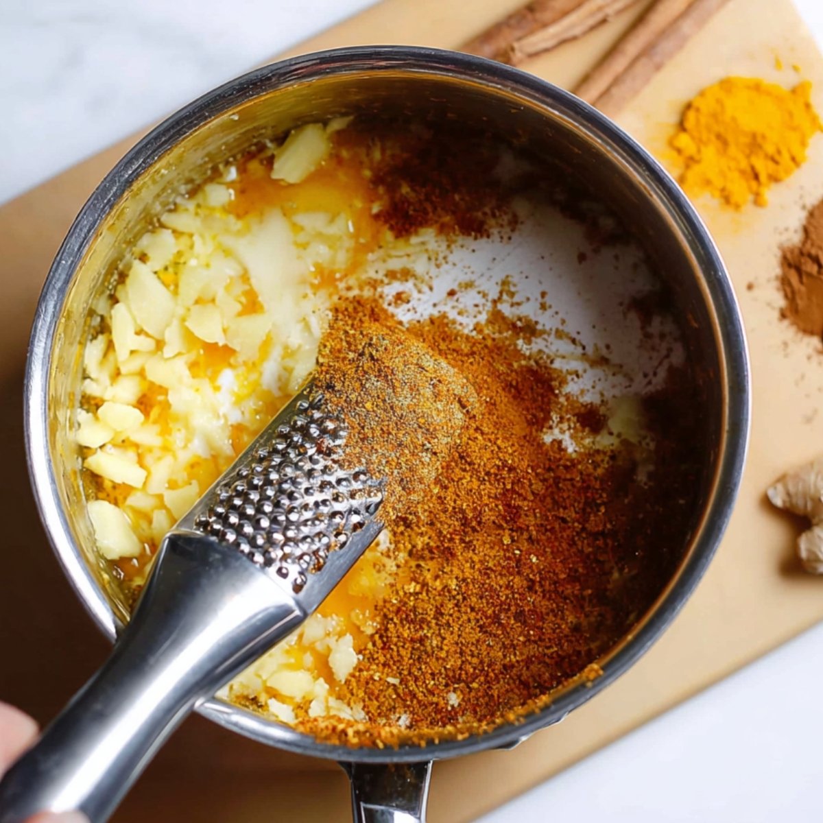"Grating fresh ginger into a saucepan with turmeric, cinnamon, and other ingredients, preparing the base for golden milk."