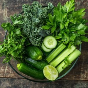 A bowl filled with fresh ingredients for green juice, including kale, cucumber, celery, parsley, and lime slices.