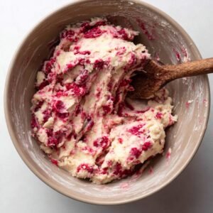 A bowl of raspberry cookie dough, with visible chunks of raspberries mixed throughout. The dough has a marbled texture and is being stirred with a wooden spoon.