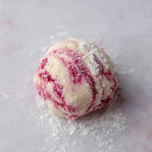 A close-up shot of a ball of raspberry cookie dough, coated with a fine dusting of flour. The dough has vibrant pink swirls from the raspberries, and it's placed on a smooth surface.