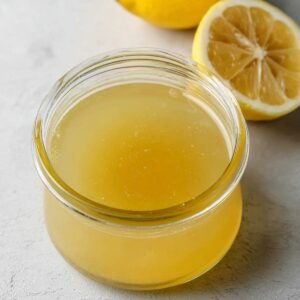 A jar of freshly squeezed lemon juice with a smooth golden color, sitting next to halved lemons. The juice is clear with small lemon pulp specks visible.