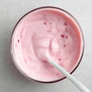 A creamy bowl of yogurt with a soft pink hue, possibly strawberry-flavored, with visible flecks of fruit inside. A white spoon is gently mixing the yogurt.