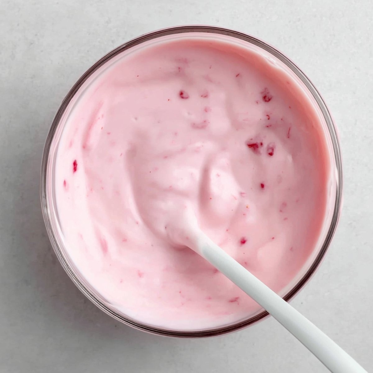 A creamy bowl of yogurt with a soft pink hue, possibly strawberry-flavored, with visible flecks of fruit inside. A white spoon is gently mixing the yogurt.