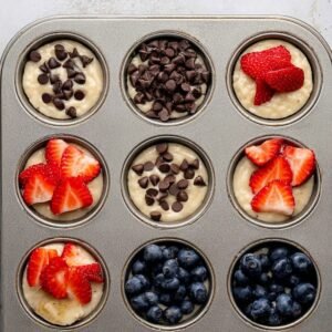 A muffin tin before baking, with muffin batter topped with fresh strawberries, blueberries, chocolate chips, and banana slices, ready to go into the oven.