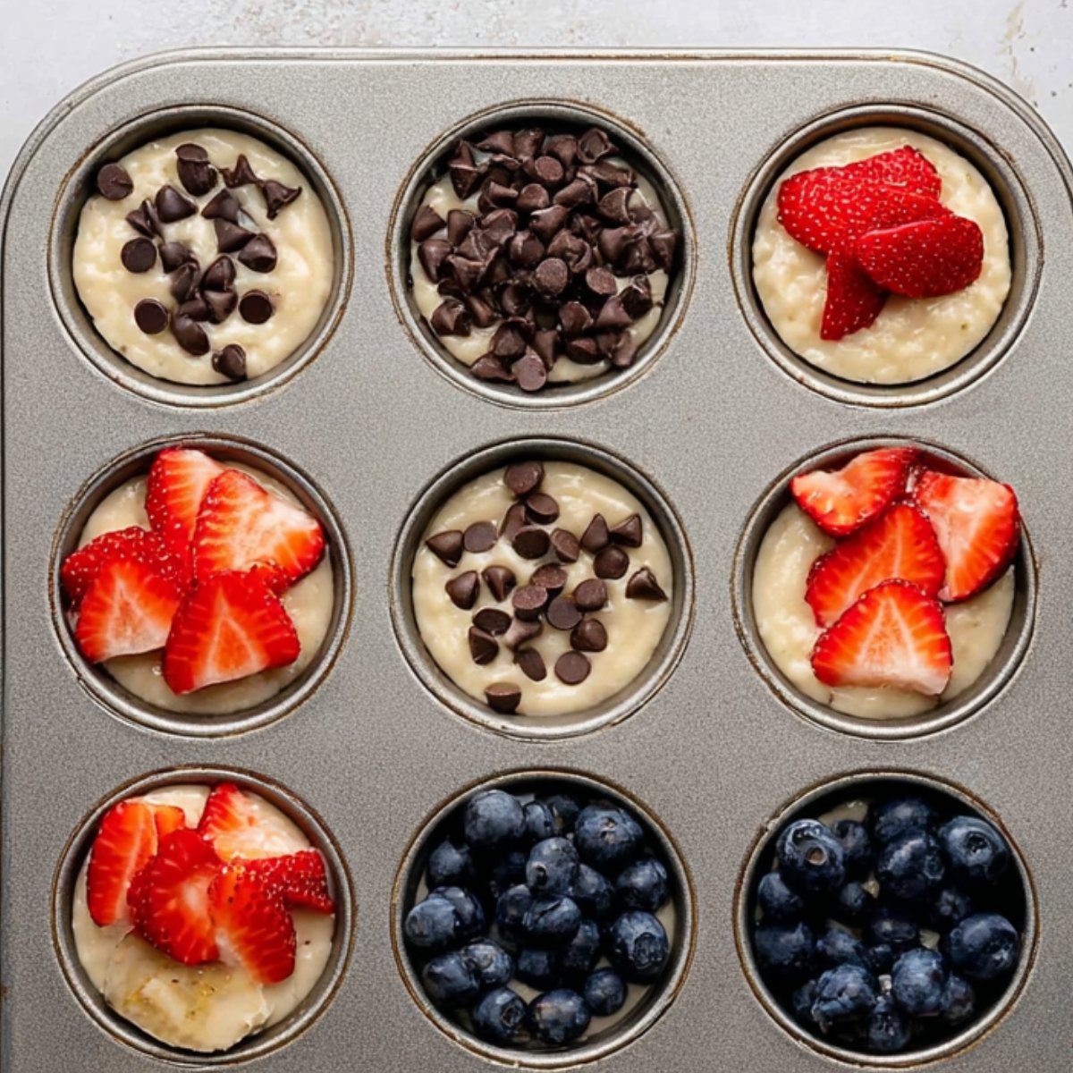 A muffin tin before baking, with muffin batter topped with fresh strawberries, blueberries, chocolate chips, and banana slices, ready to go into the oven.