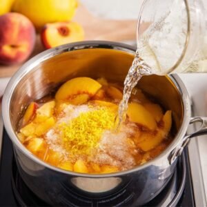 A pot on the stovetop with peaches, lemon zest, and sugar, being prepared for a peach lemonade mix. A water jug is pouring liquid into the pot while lemons and peaches sit nearby.