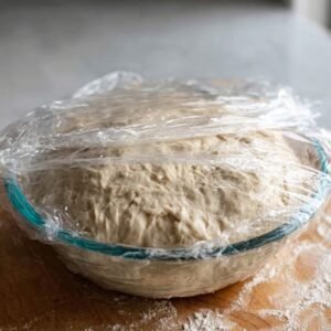 "A person covers a bowl of bread dough with plastic wrap to let it rise."