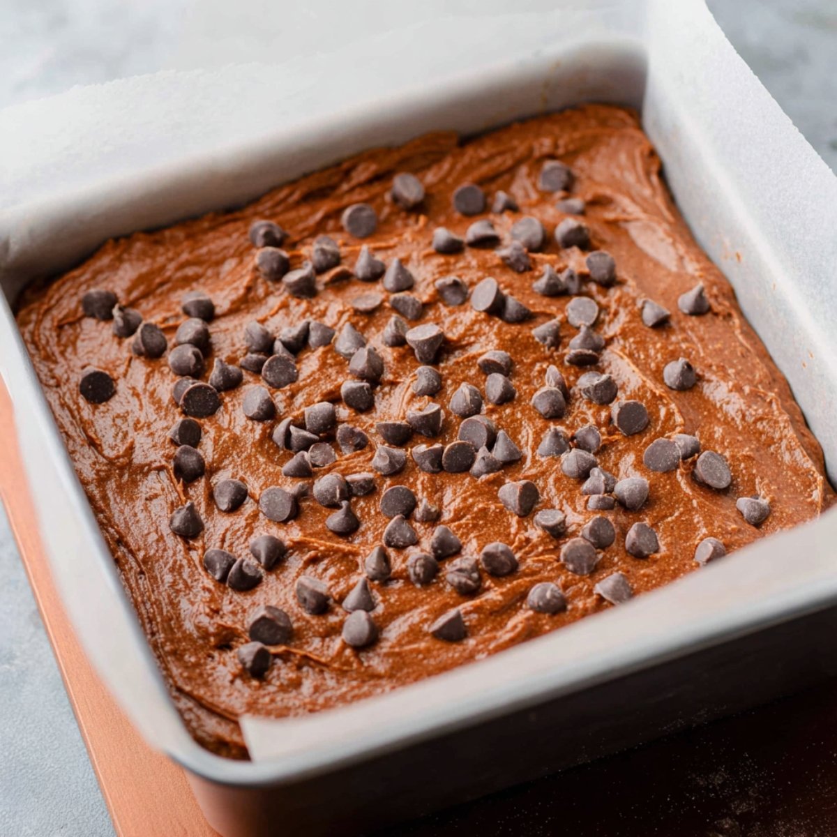 "A close-up shot of the brownie batter spread in a baking pan, topped with chocolate chips, ready to be baked.