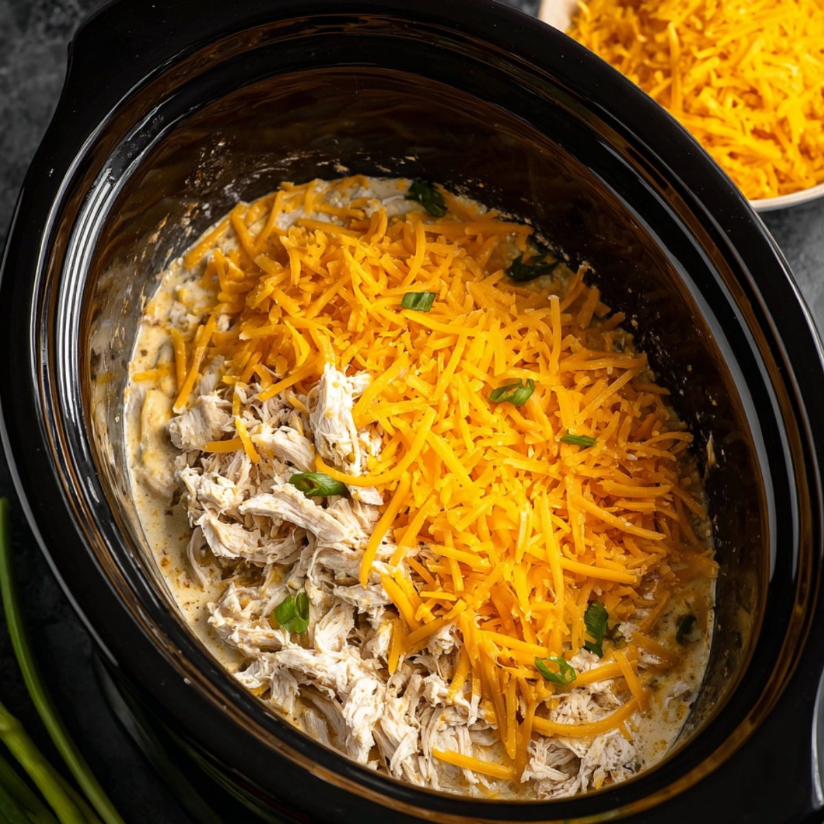 Overhead view of shredded slow cooker chicken partially covered with shredded cheddar cheese and scattered chopped green onions, creamy sauce visible underneath, in a black slow cooker, ready to be mixed and served.