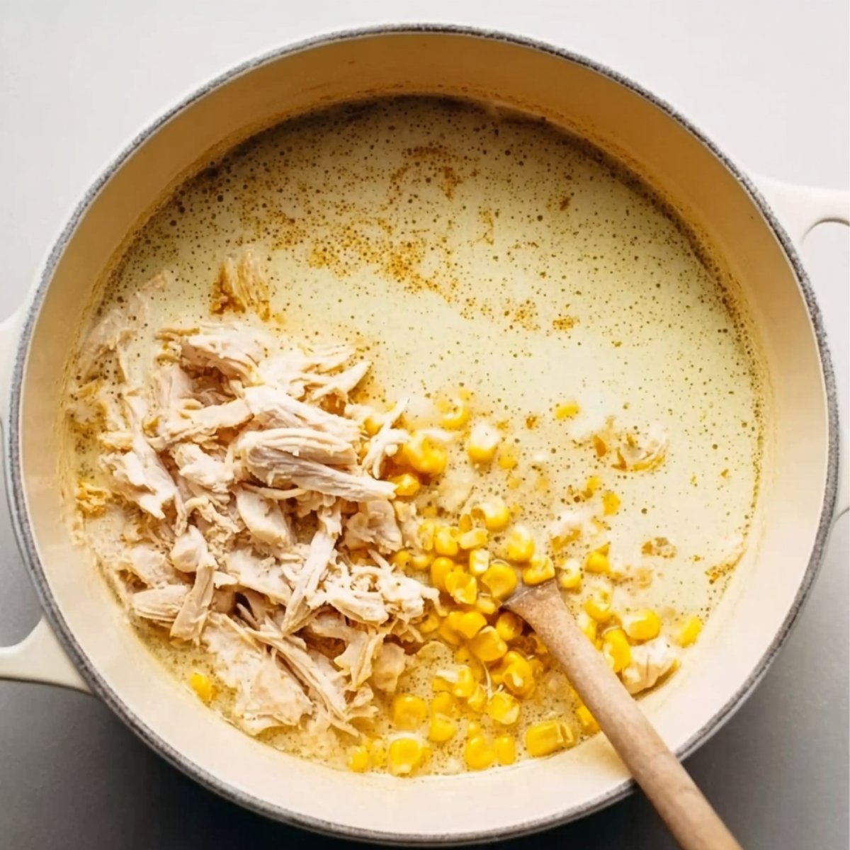 Top-down view of a pot with creamy soup base, shredded chicken, and yellow corn added, ready to be stirred, with a wooden spoon partially visible.