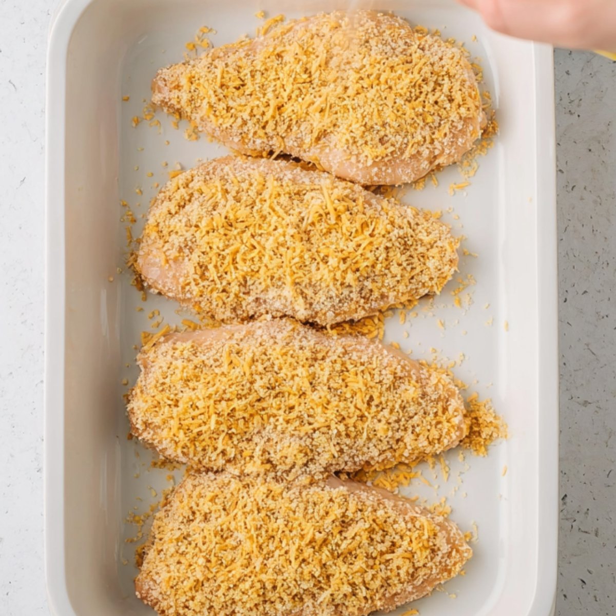 aw chicken breasts in a white rectangular baking dish, coated with a cheesy mixture and sprinkled with panko breadcrumbs, ready for baking.