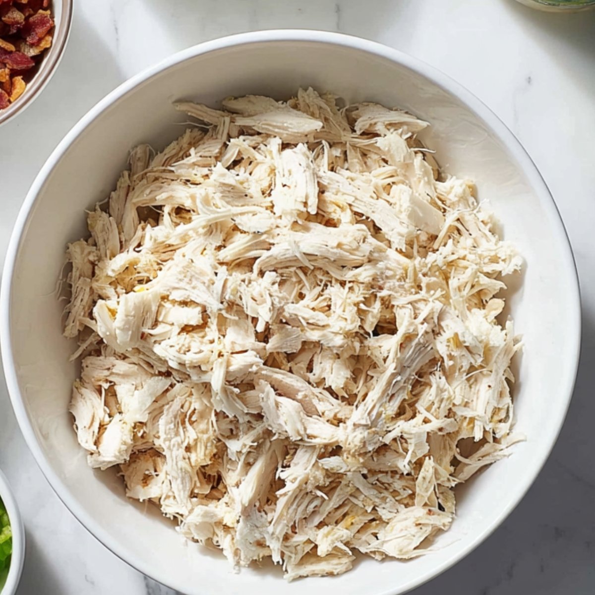 A white bowl filled with shredded cooked chicken, ready for use in recipes like quesadillas or tacos. Surrounding the bowl are partial views of other ingredients, including chopped green onions and dried tomatoes.