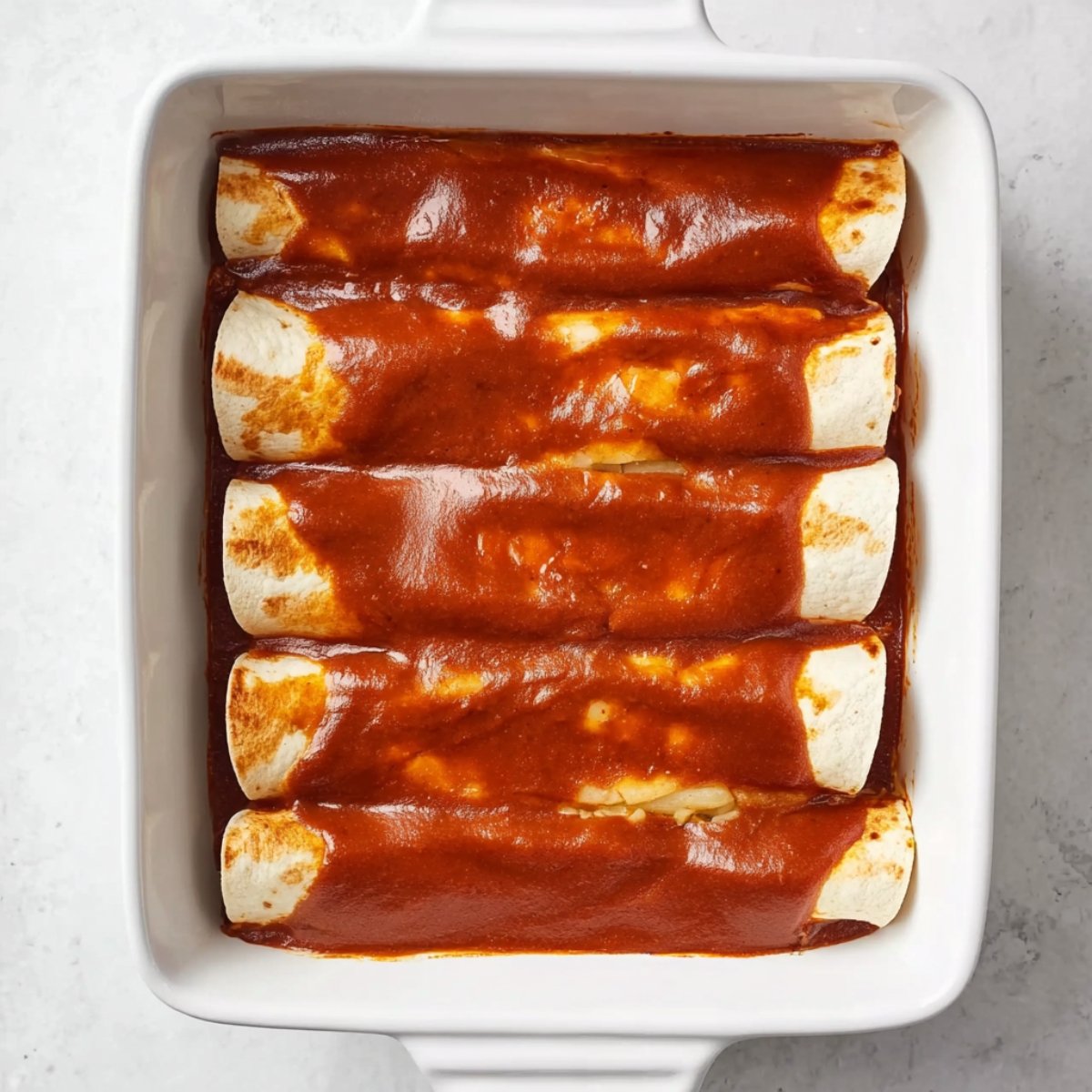 Top-down view of fully assembled beef and pineapple enchiladas in a white rectangular baking dish, covered with red enchilada sauce, drizzled with sour cream, and topped with pineapple chunks and chopped cilantro, black spatula and green kitchen towel beside the dish, bright natural lighting.